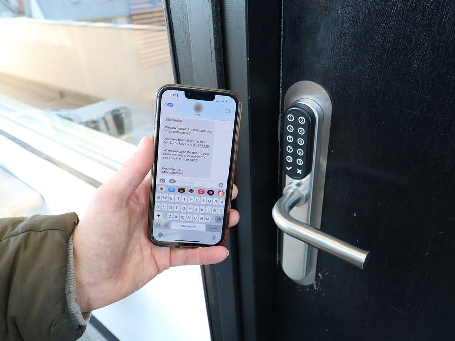 Eine Hand hält ein Smartphone, auf dem eine Nachricht zur Zimmerzuweisung angezeigt wird, und ein Tür mit einem Zahlenschloss ist zu sehen.