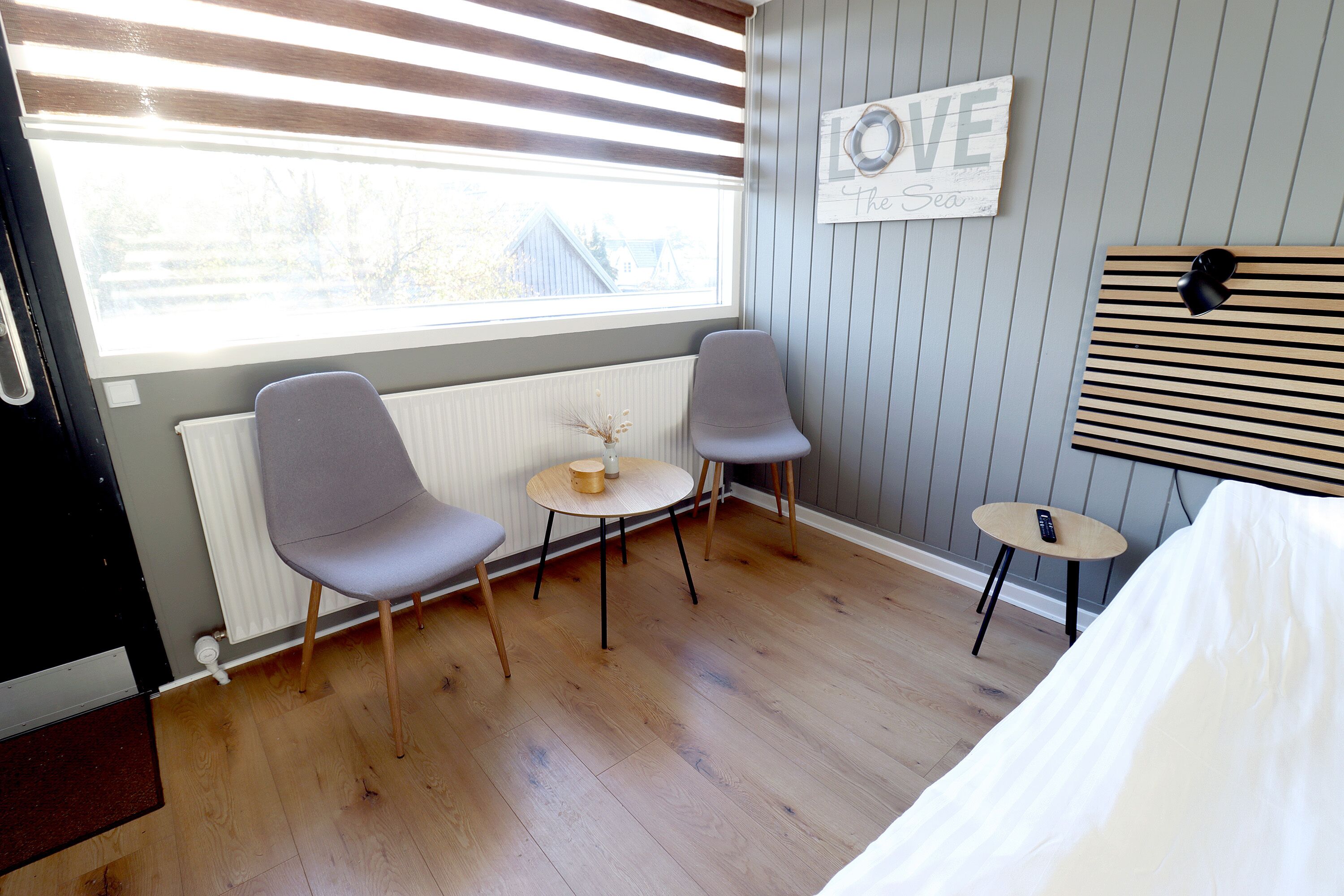 Quarto com cama, duas cadeiras, uma mesinha, radiador, janela com persianas e um painel na parede com a inscrição 'LOVE'.