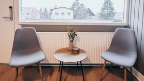Una habitación con dos sillas grises, una mesa redonda pequeña con un florero, y un ventanal con vistas a edificios y árboles.