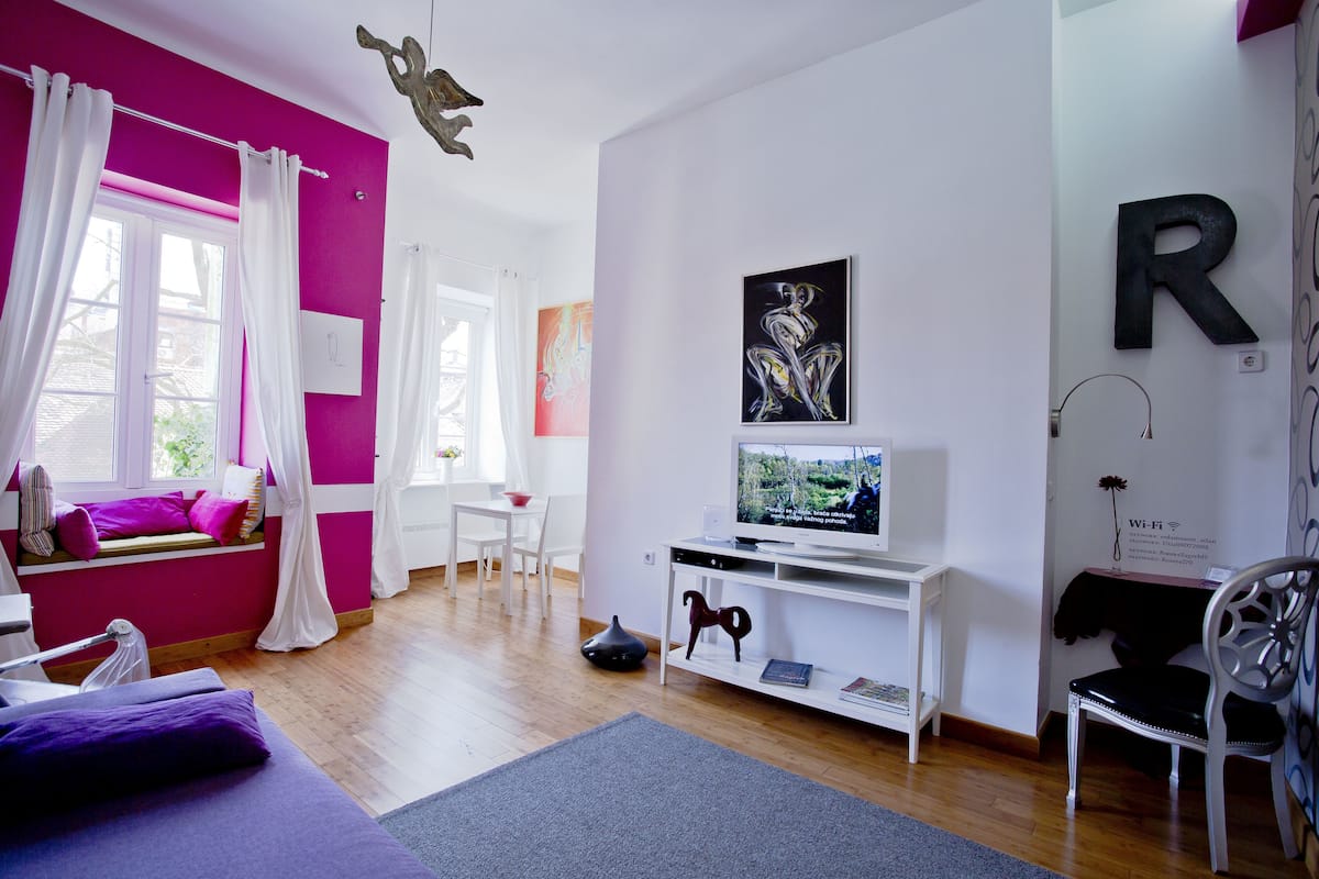 Ein modernes Wohnzimmer mit einer leuchtend pinkfarbenen Akzentwand, einer weißen Unterhaltungsanlage und einer Fensterbank mit violetten Kissen.