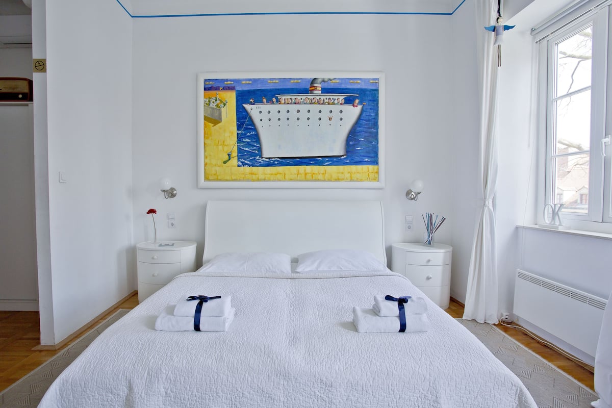 Ein Hotelzimmer mit einem großen Bett, zwei Nachttischen, einem gerahmten Kunstwerk über dem Bett und einem Fenster zur rechten Seite.