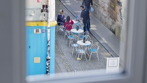 Vista a través de una ventana que muestra una calle con mesas al aire libre, un pilar azul y un edificio con grafitis.