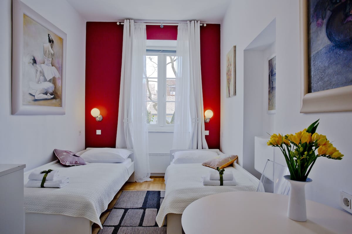 Ein Hotelzimmer mit zwei Betten, einem Fenster mit weißen Vorhängen, einer Vase mit gelben Blumen und Bildern an den Wänden.