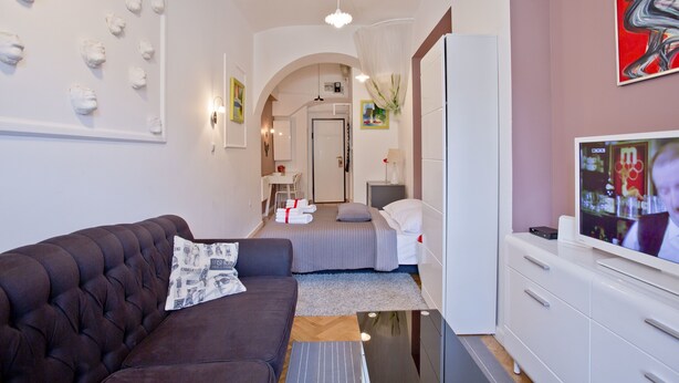 Una habitación de hotel moderna con un sofá marrón oscuro, un televisor de pantalla plana, un armario blanco y una cama con ropa de cama gris.