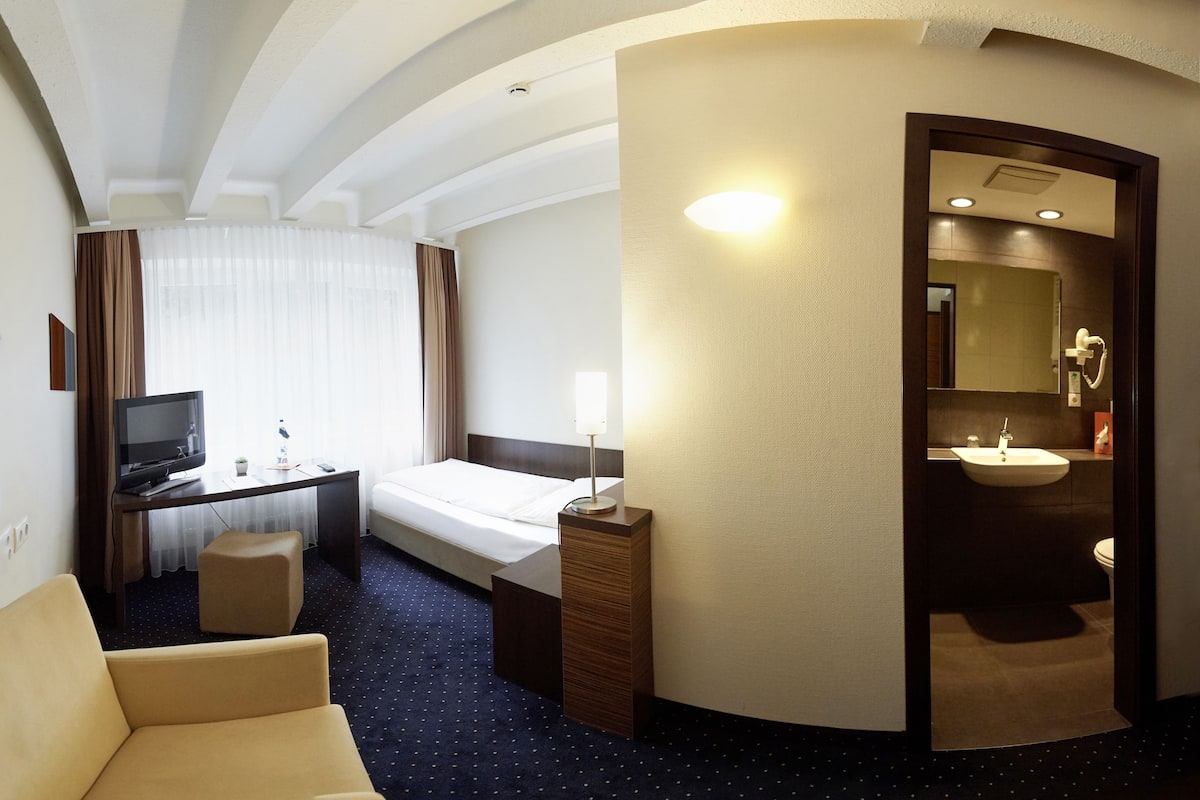 Habitación de hotel con cama, escritorio con computadora, silla, mesita y baño con lavamanos y espejo.