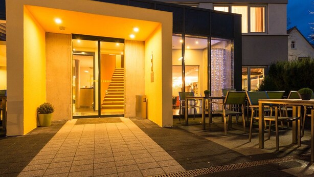 A modern building with a sign reading "Silcium Restaurant Location Hotel." It features a welcoming entrance with a staircase, outdoor seating area, and a well-lit facade.
