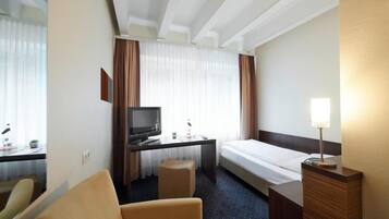 A hotel room with a bed, a desk with a television, a chair, a bedside table with a lamp, and a window with curtains.