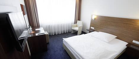 Una habitación de hotel con cama, escritorio, silla, televisor y ventana con cortinas.