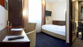 A hotel room with a bed, a desk with a remote control, a chair, a TV mounted on the wall, and a bathroom visible through an open door.