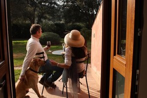 Un homme et une femme sont assis à une table à l’extérieur, accompagnés d’un chien, dans un patio donnant sur un jardin.