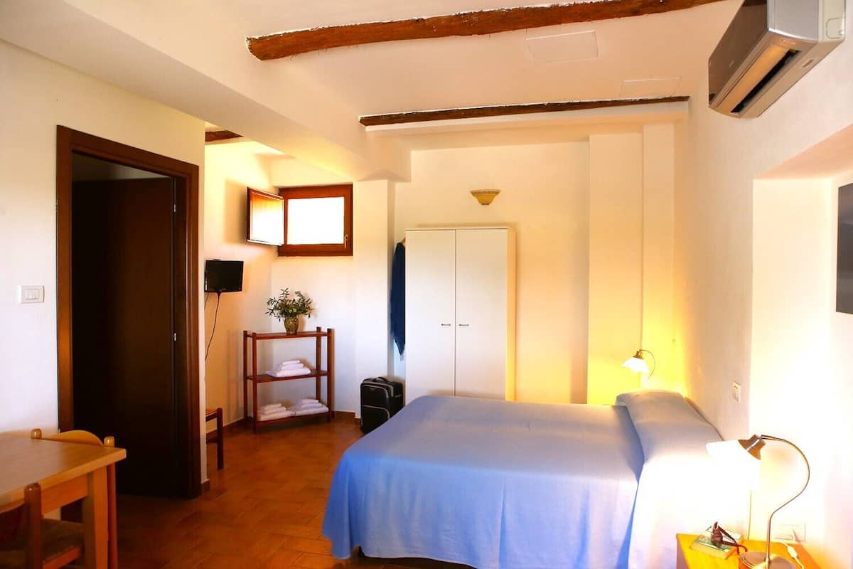 A hotel room with a bed, a desk, a chair, a wardrobe, and a small table with a lamp.