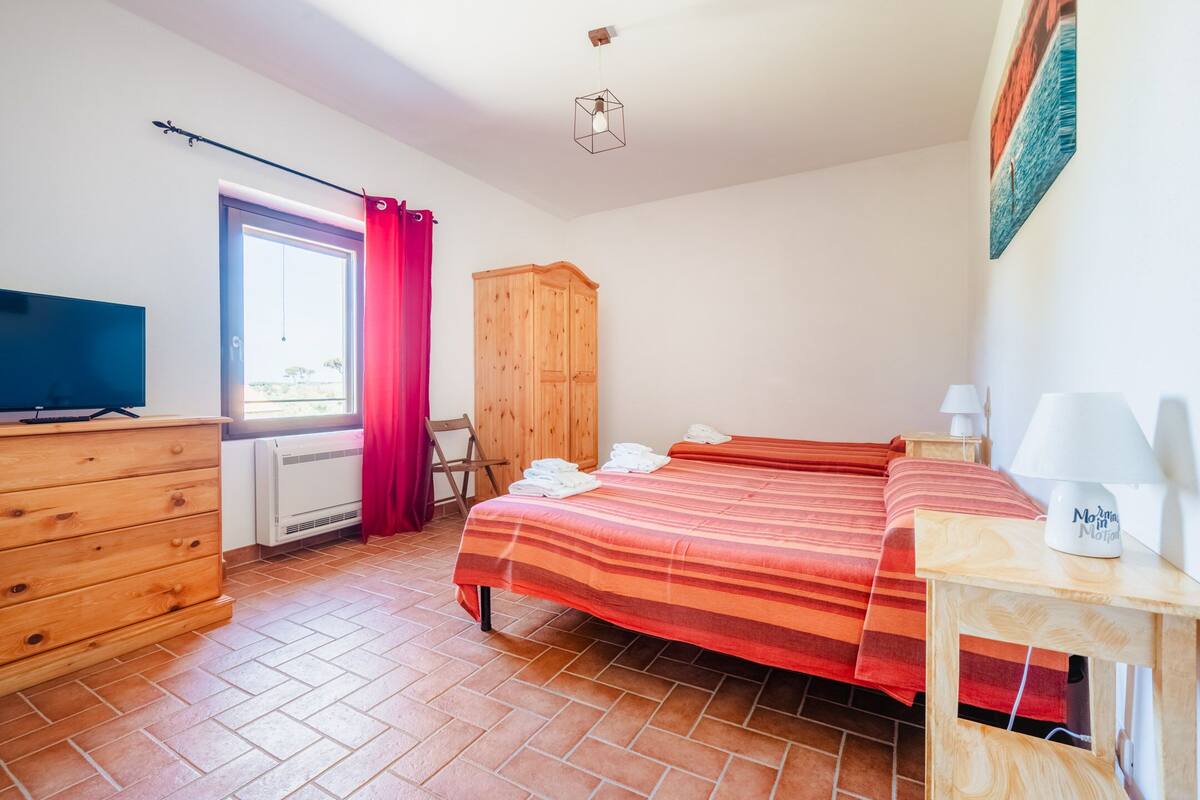 A bedroom with a bed, a wooden wardrobe, a dresser, a desk with a lamp, and a television.