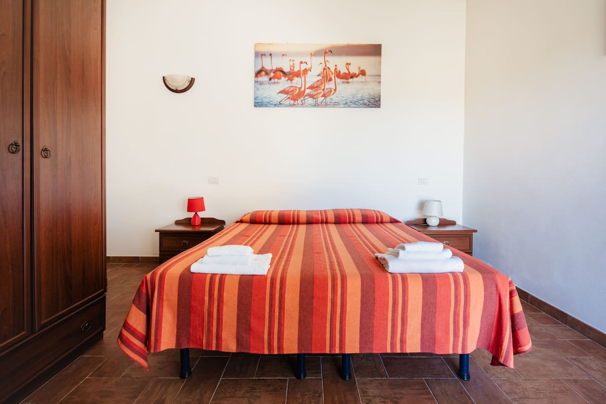 A hotel room with a large bed, a wooden wardrobe, a nightstand with a red lamp, and a painting of flamingos on the wall.