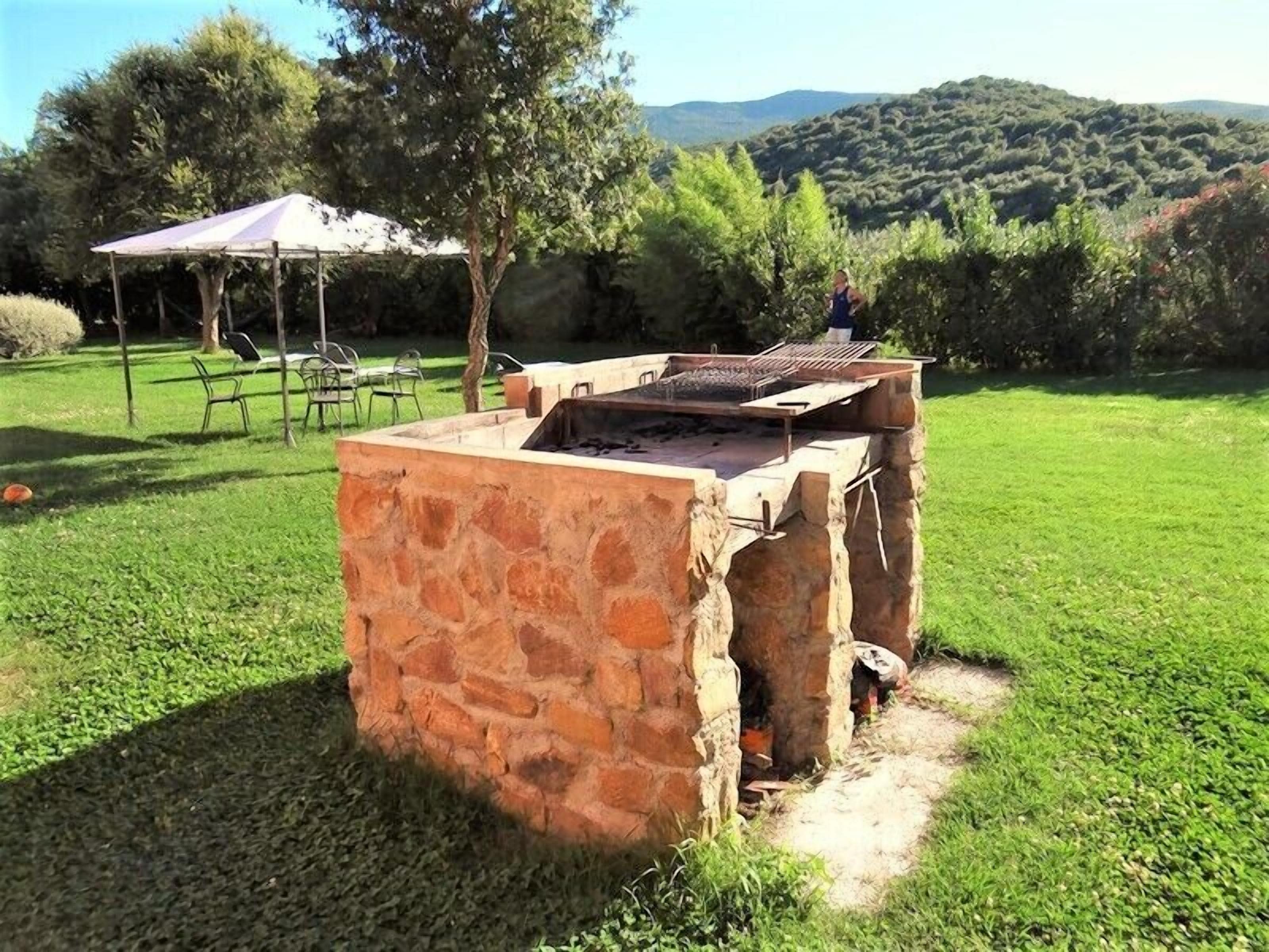 Una parrilla de piedra para barbacoa al aire libre en un césped, con una mesa de picnic y una sombrilla al fondo.