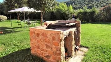 Una parrilla de piedra para barbacoa al aire libre en un césped, con una mesa de picnic y una sombrilla al fondo.