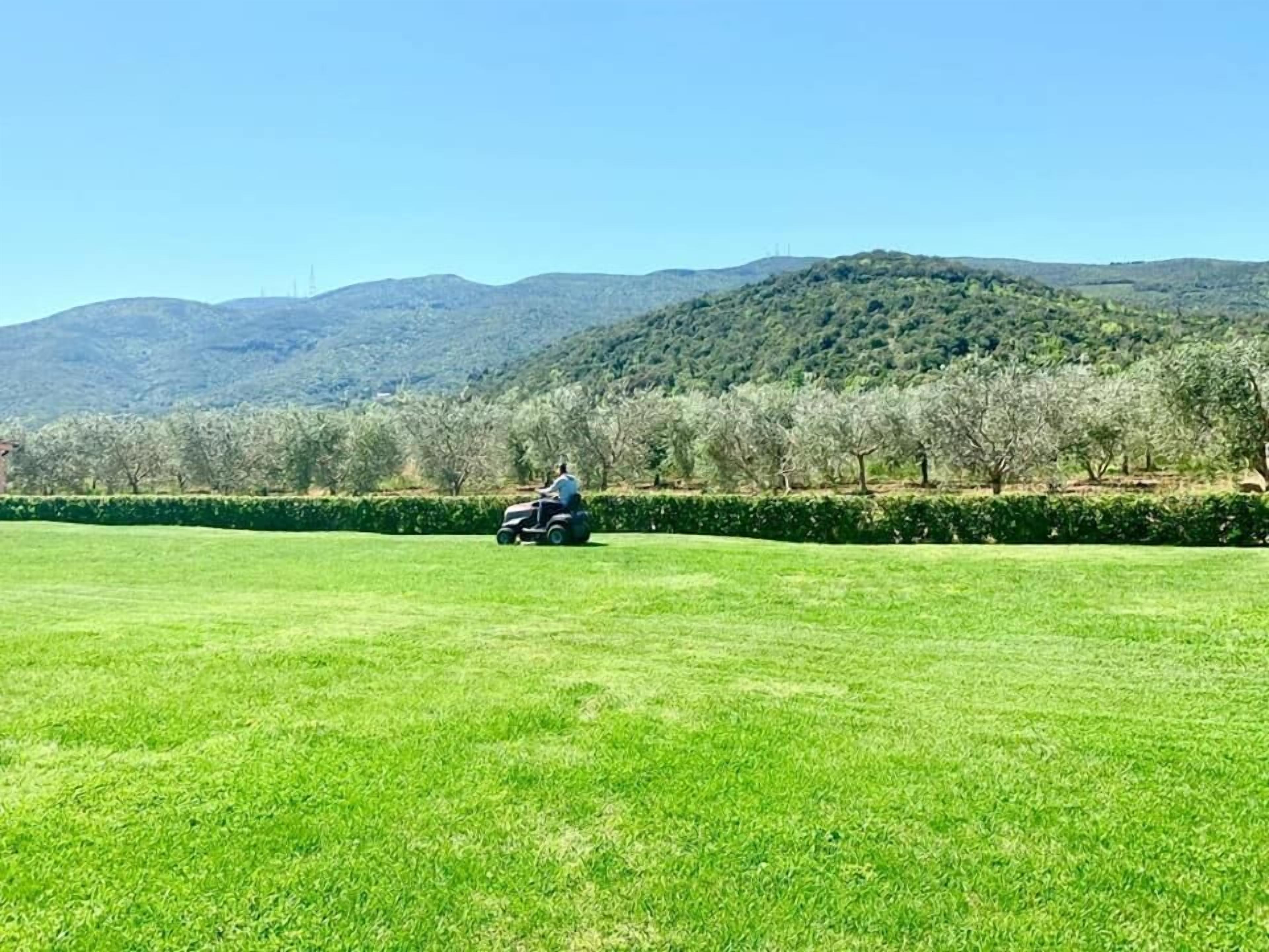 A person riding a lawn mower on a green field with trees and hills in the background.