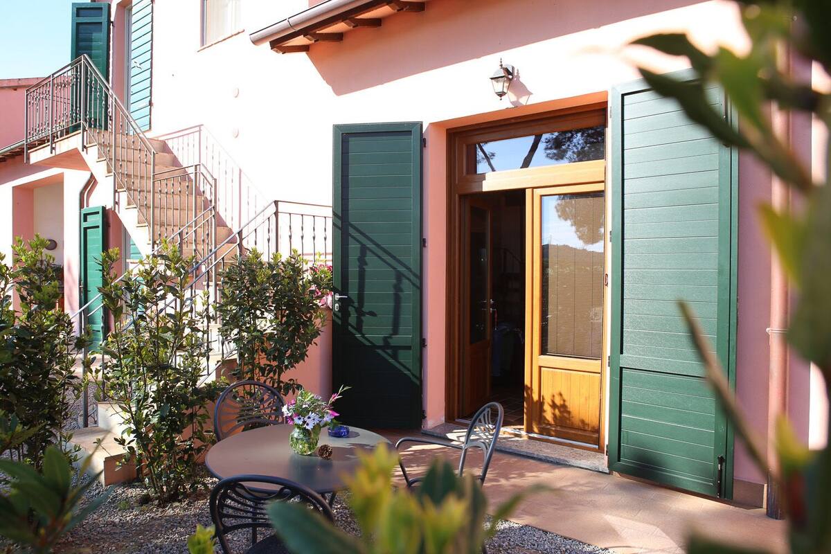 A patio with a table and chairs, a small garden, and a building with green shutters and a wooden door.