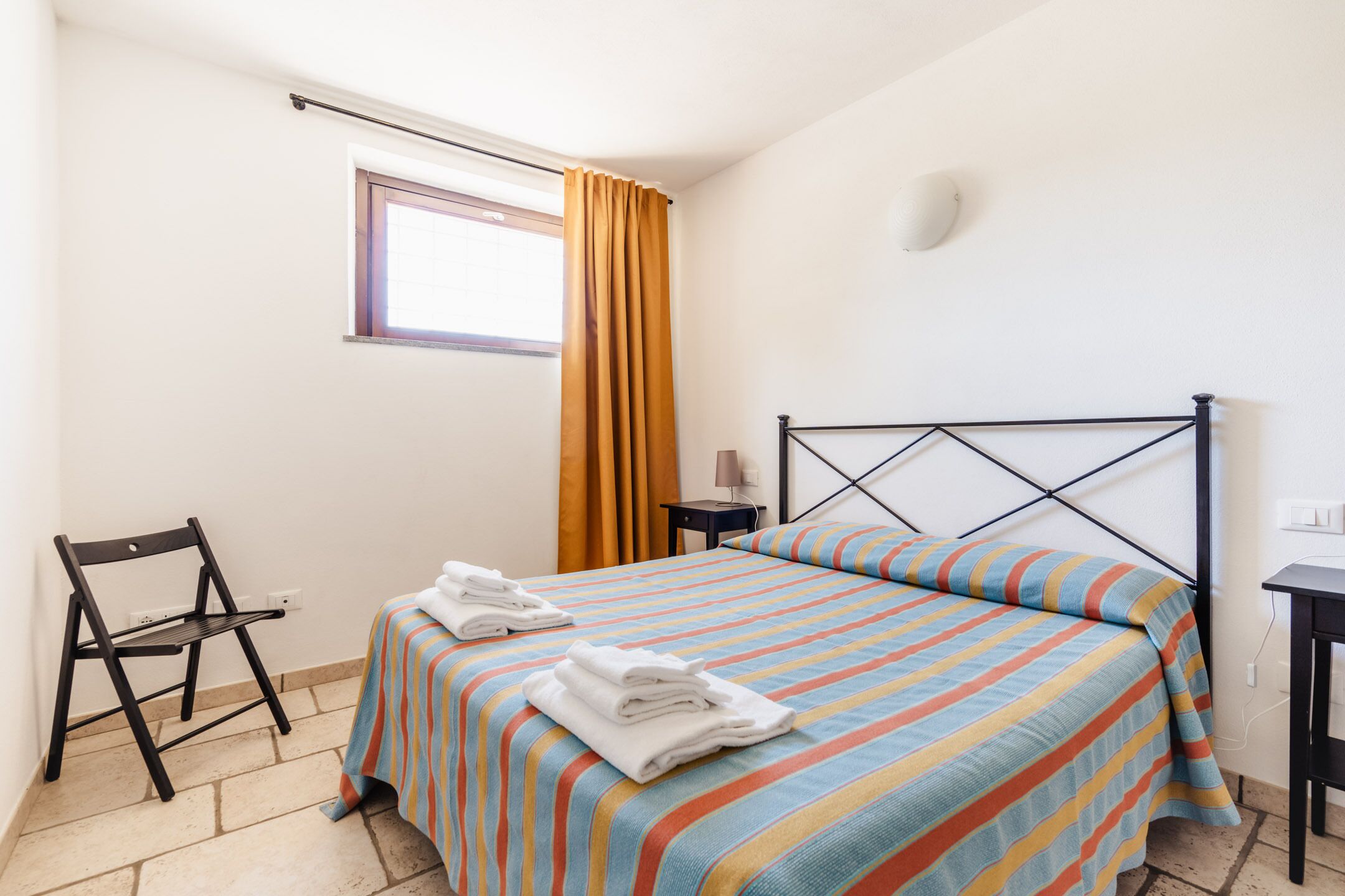 Una cama bien hecha con ropa de cama a rayas, una silla, una mesita con una lámpara y una ventana con cortinas.