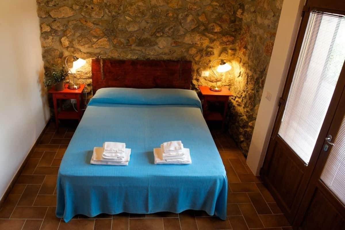 A room with a stone wall, a bed with a blue bedspread, two bedside tables with lamps, and a door with blinds.