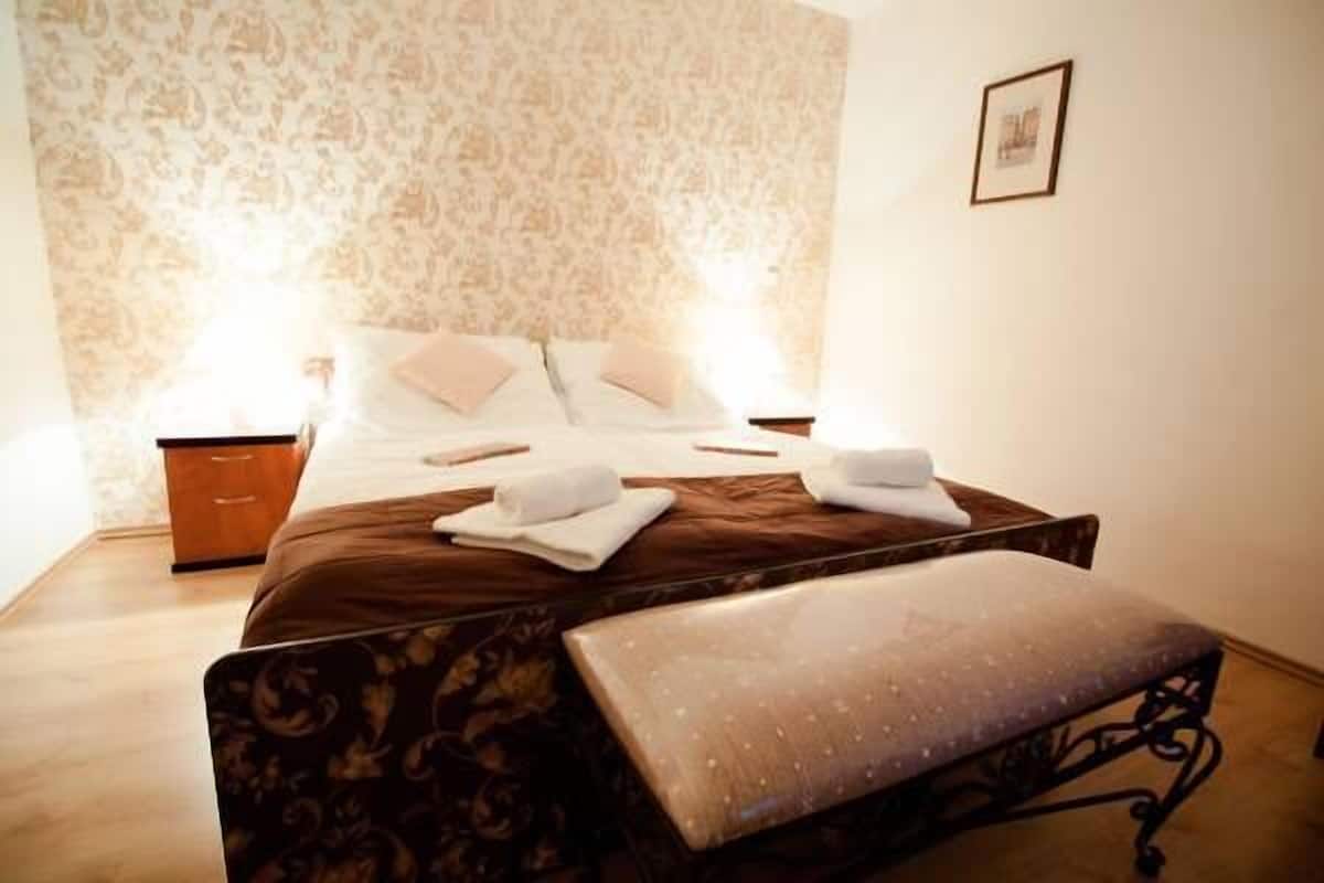 Ein ordentlich bezogenes Bett mit weißen Leinen und einem braunen Kopfteil, ein Nachttisch mit Lampe sowie ein kleines gerahmtes Bild an der Wand.