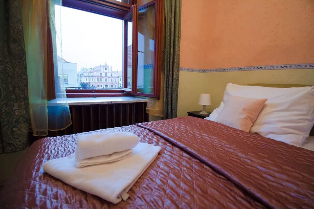 Ein Hotelzimmer mit Bett, Fenster mit Blick auf Gebäude, Nachttisch mit Lampe und Vorhängen.