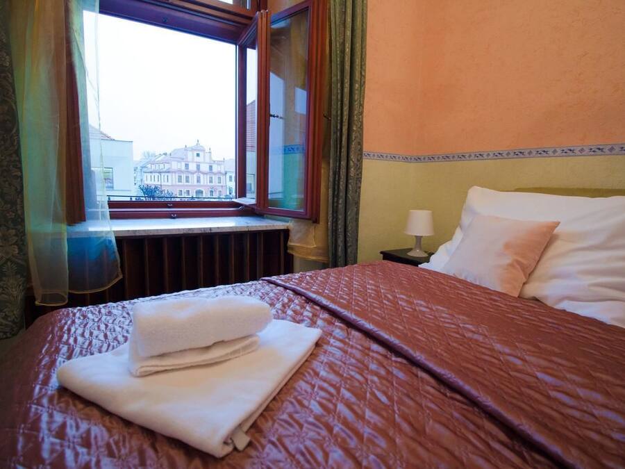 A hotel room with a bed, a window with a view of buildings, a nightstand with a lamp, and a window with curtains.