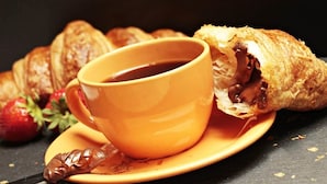 Een kop koffie op een schotel met een croissant en een chocoladegebakje op een bord.