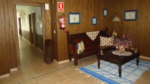 A wooden-finished hallway with a fire extinguisher mounted on the wall, a sofa with patterned cushions, a coffee table with a vase of flowers, and framed pictures on the wall. - As Maceiras (Puebla de Trives)