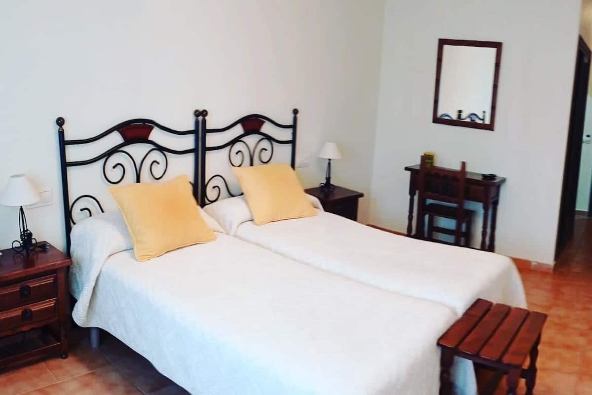 A double bed with a white bedspread and two pillows, a wooden nightstand with a lamp, a wooden bench, and a small table with a lamp.