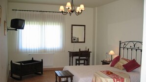 A bedroom with a bed, bedside table, chair, and a small table. There is a window with curtains, a radiator, a television on the wall, and a chandelier. - As Maceiras (Puebla de Trives)