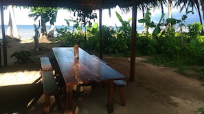 An outdoor wooden table and benches under a thatched roof, overlooking a beach with palm trees and the sea.