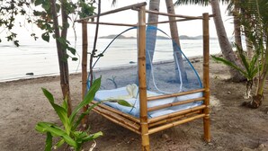 Una poltrona a dondolo in bambù con cuscino bianco e zanzariera blu, posizionata su una spiaggia sabbiosa con palme e vista sul mare.