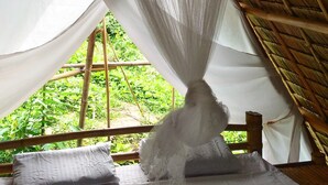 Una stanza con un letto sotto un tetto di paglia, una zanzariera e vista sul verde circostante.