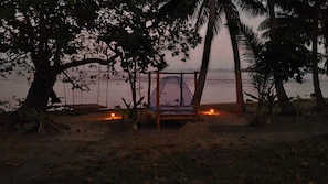 A beach scene with a wooden bed under a tree, a small fire, and a tent.