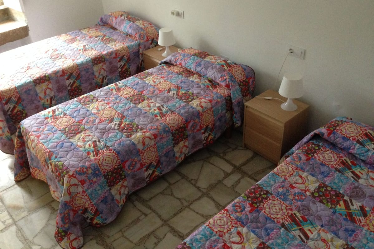 A room with three single beds, each with a colorful quilt. There are two bedside tables with lamps, and the room has a tiled floor.