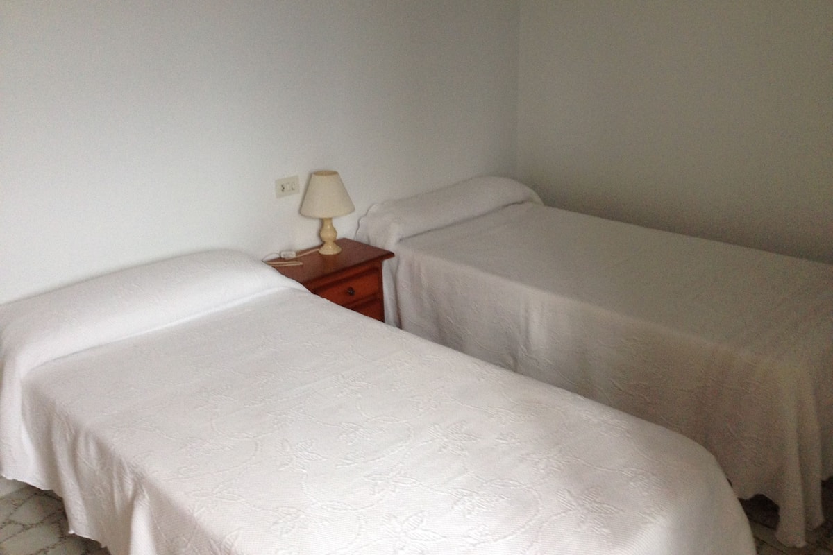 A small, simple room with two single beds, a nightstand with a lamp, and a plain white wall.
