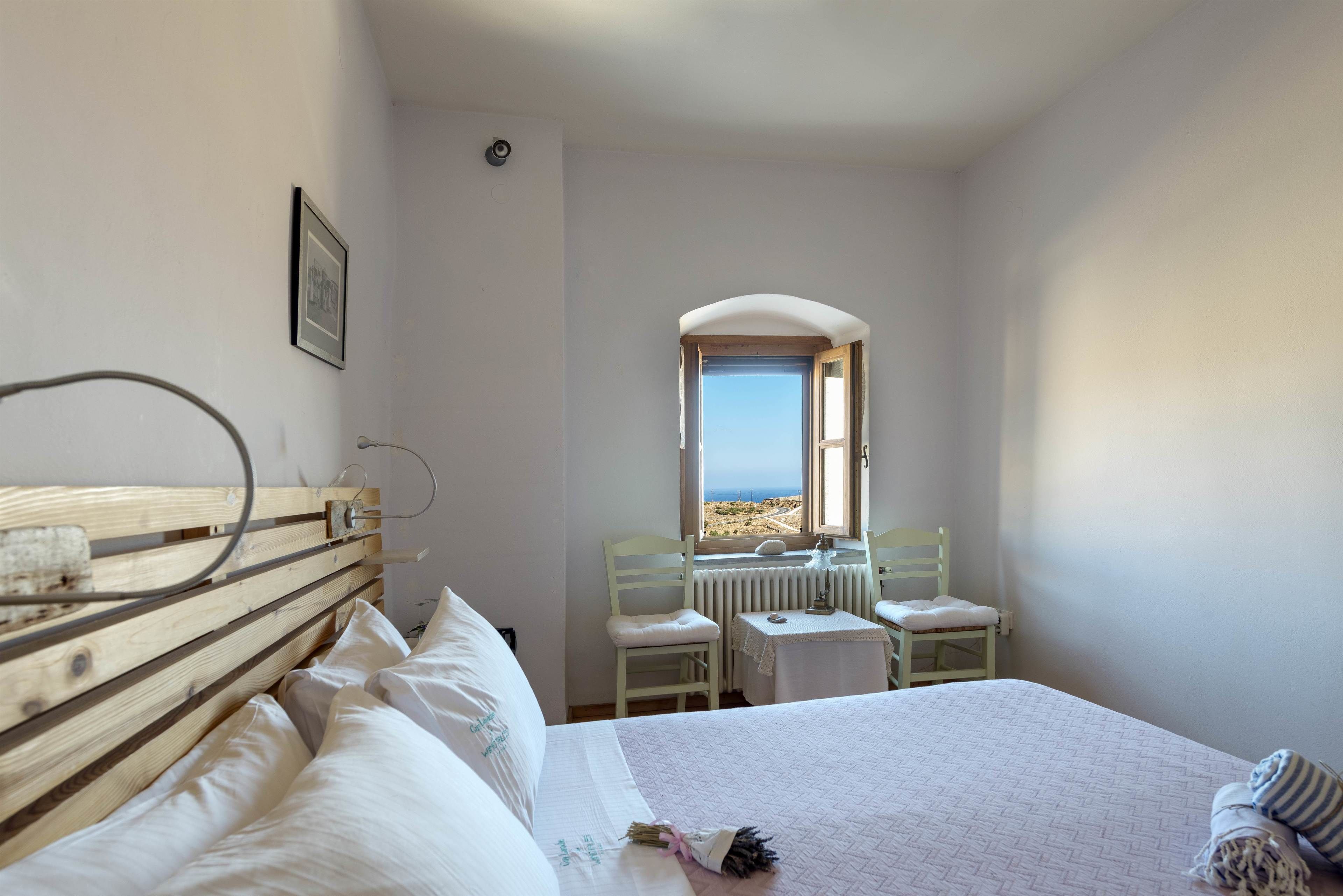Un dormitorio con cabecera de madera, una cama con ropa de cama blanca, una pequeña mesa con una silla y vista al mar a través de una ventana.