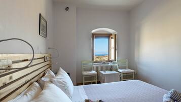 Un dormitorio con cabecera de madera, una cama con ropa de cama blanca, una pequeña mesa con una silla y vista al mar a través de una ventana.