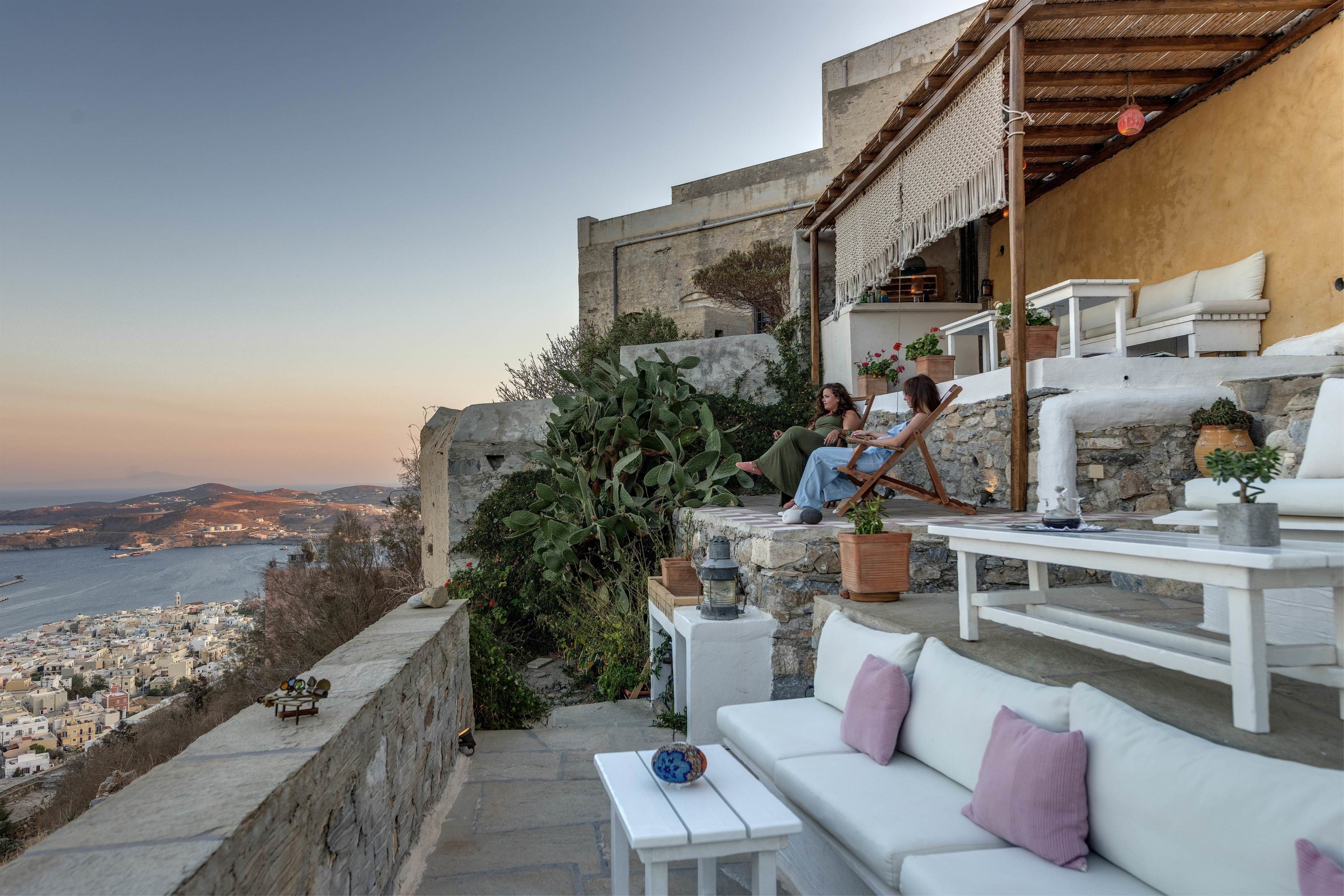 A terrace with white furniture, a stone wall, and a view of a coastal city.