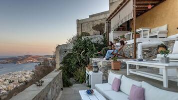 A terrace with white furniture, a stone wall, and a view of a coastal city.