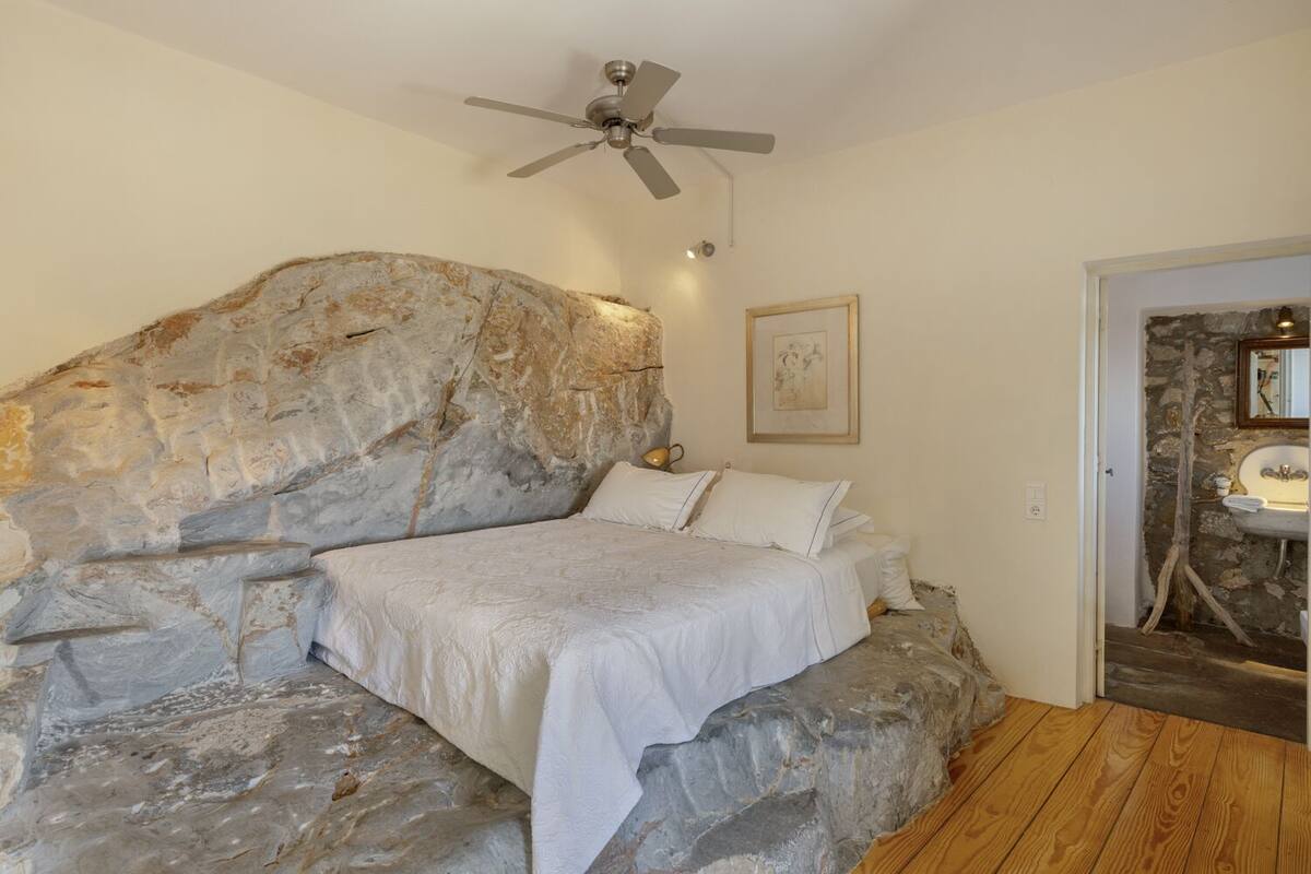 Un dormitorio con cabecera de piedra, cama con ropa de cama blanca, ventilador de techo y baño rústico a la vista a través de una puerta abierta.