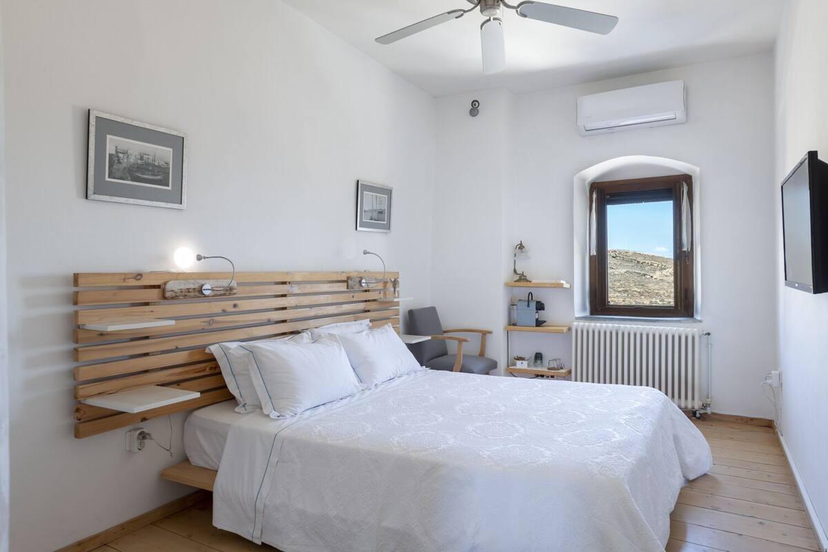 Un dormitorio con cabecera de madera, cama con ropa de cama blanca, escritorio con silla, ventana con vista y ventilador de techo.