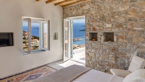 A bedroom with a stone wall, a bed, a TV, and a view of the sea.