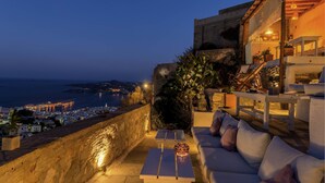 A terrace with a white sofa and a small table, overlooking a coastal cityscape at dusk.