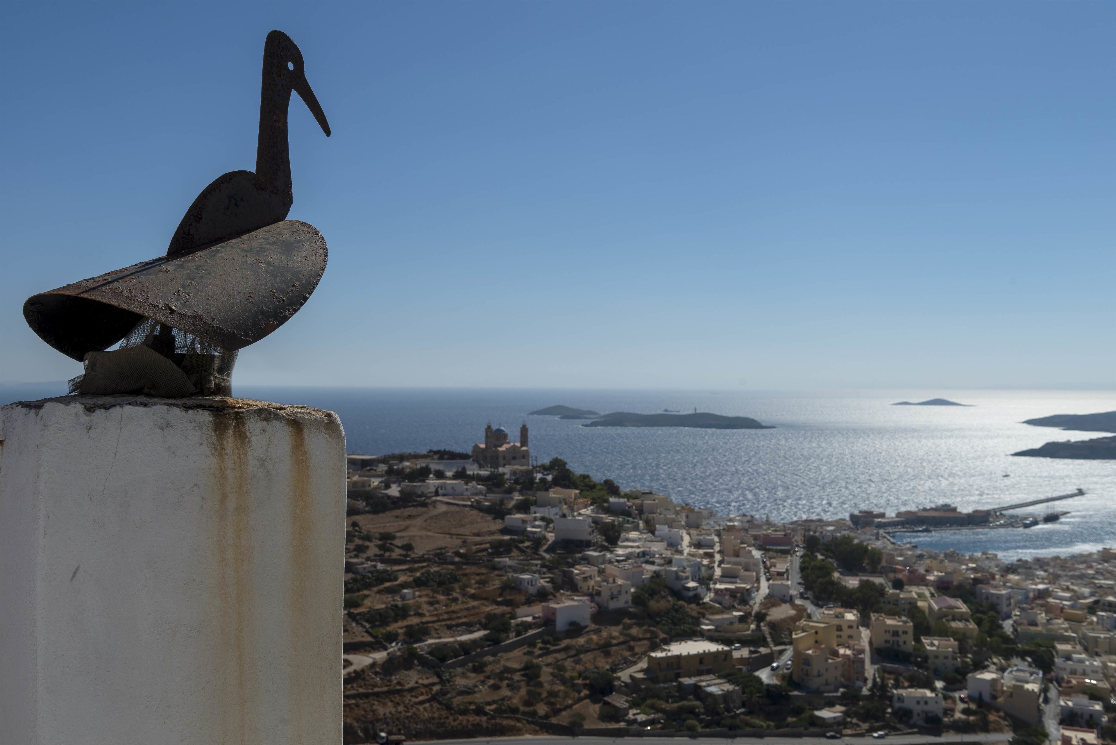 A coastal town with a prominent bird sculpture on a white pillar, overlooking a harbor and distant islands.