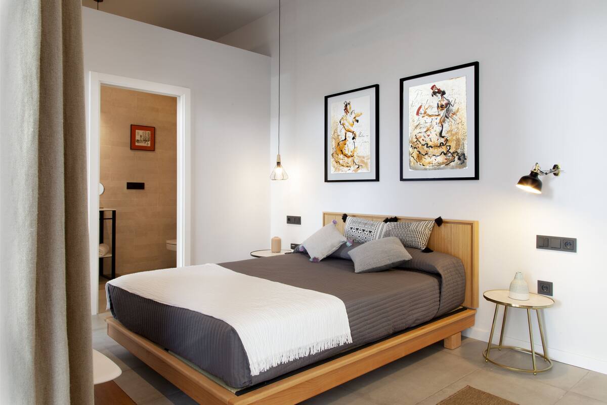 A modern bedroom with a wooden bed, a bedside table, a wall-mounted lamp, and two framed artworks.