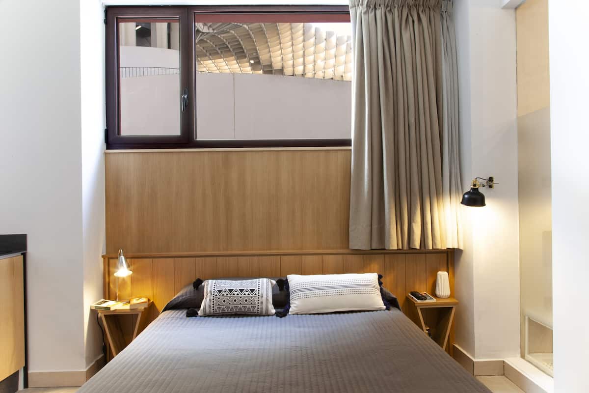 A hotel room with a large bed, wooden headboard, two bedside tables with lamps, a window with curtains, and a view of a building outside.