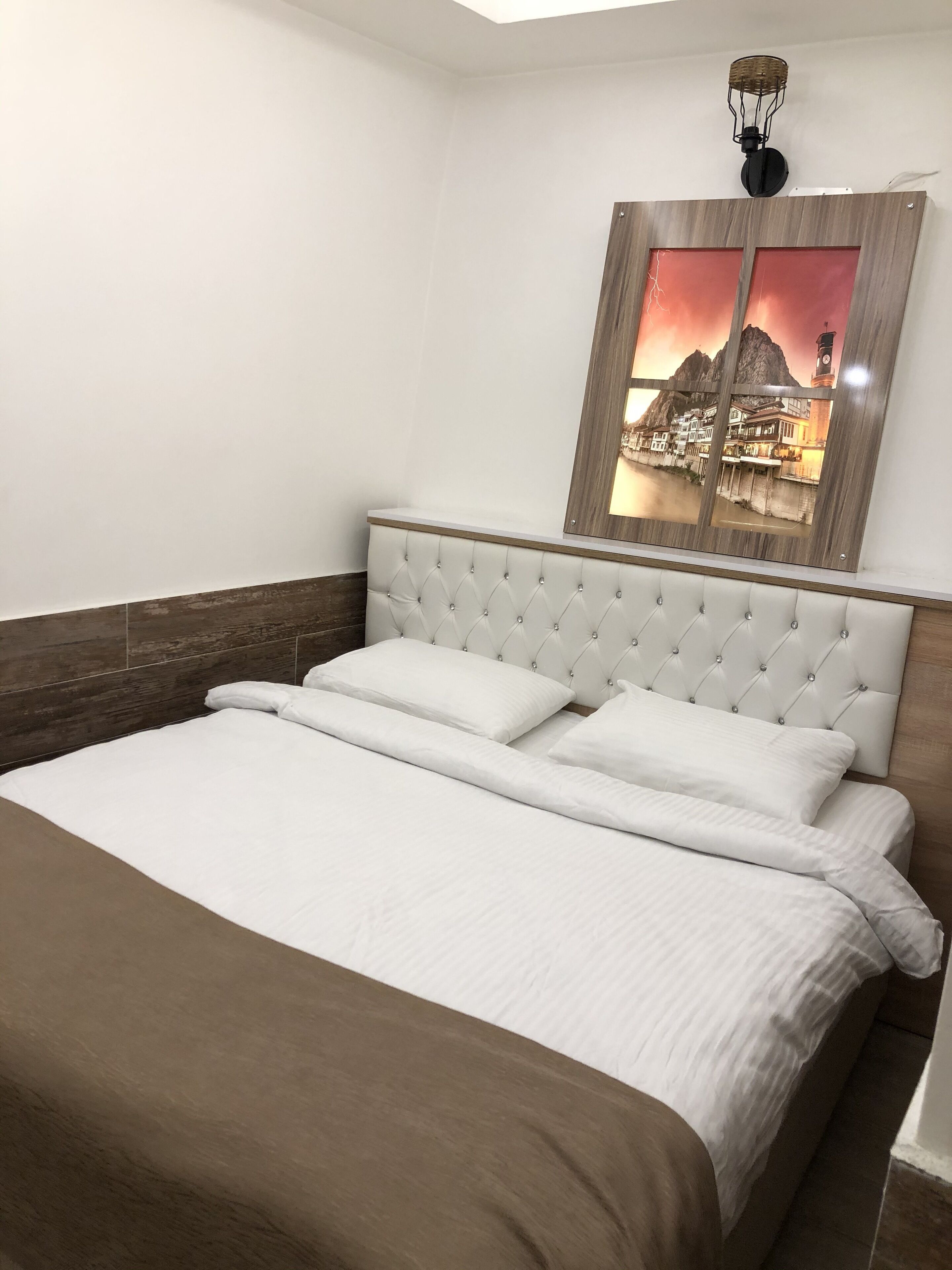 Een net opgemaakt bed met witte lakens en een bruine plaid, een gestapeld hoofdbord en een aan de muur hangend beeld van een kustplaats.