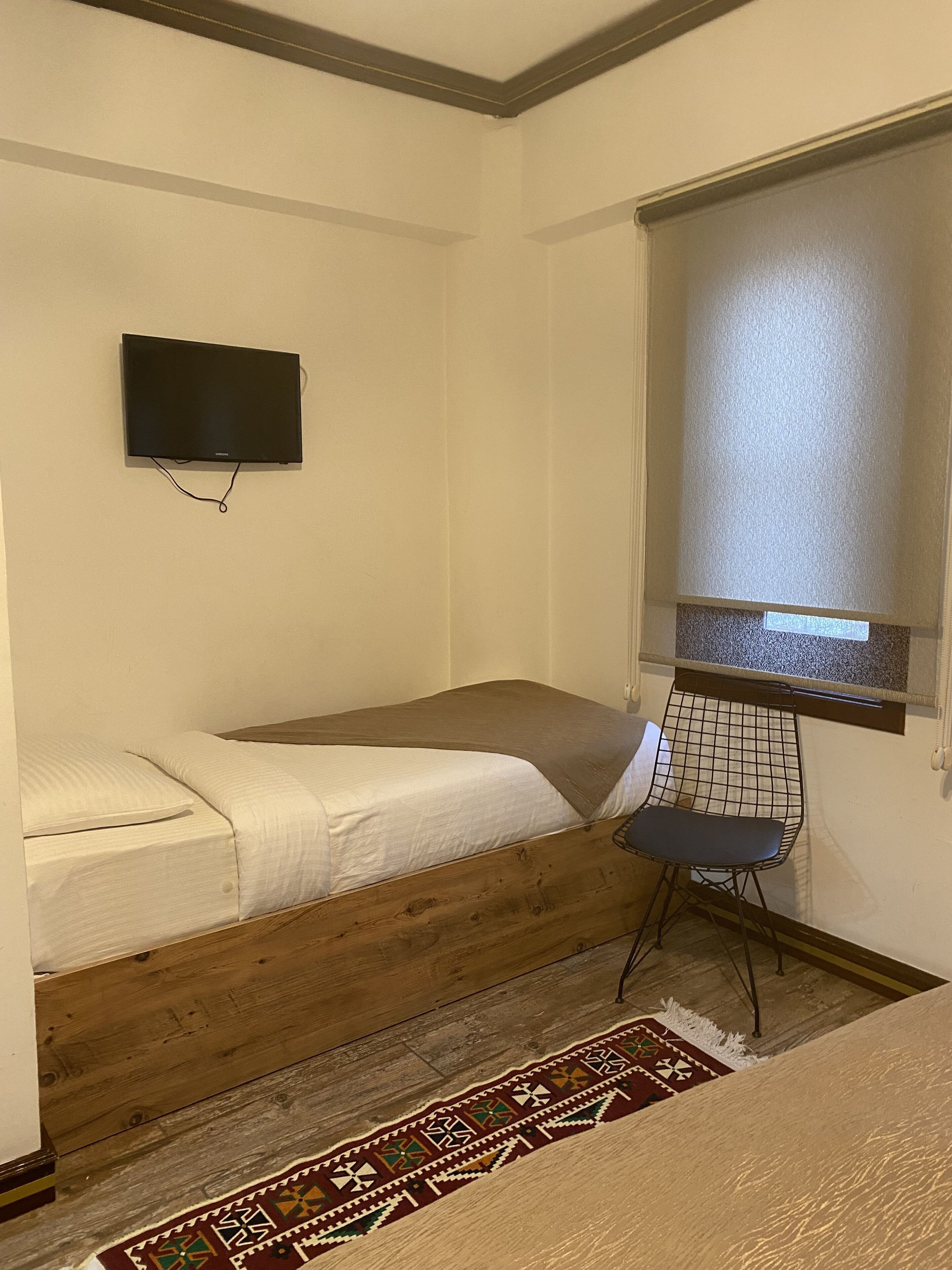 A small, sparsely furnished room with a single bed, a chair, a television mounted on the wall, and a window with blinds.