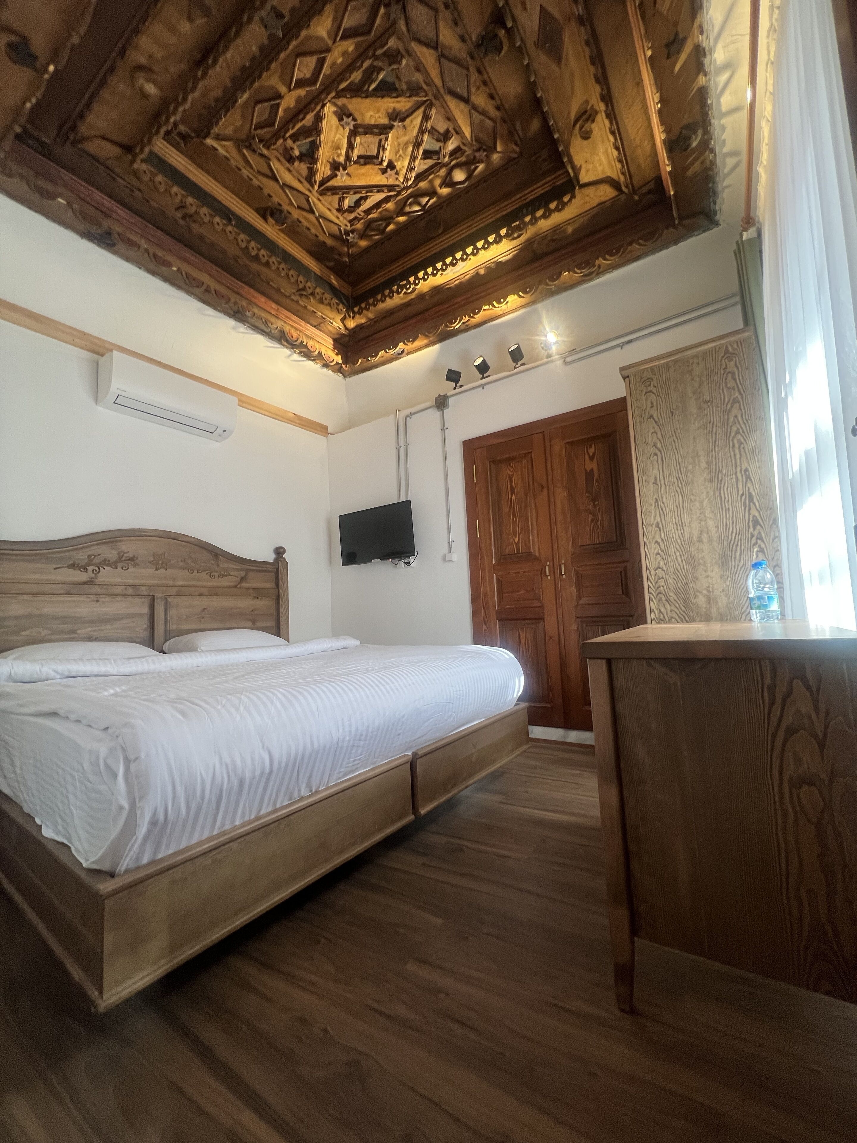 A bedroom with a wooden bed, a wooden desk, a television mounted on the wall, and a wooden ceiling with intricate designs.